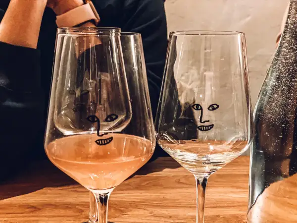 Dwa kieliszki z zabawnymi buźkami na drewnianym stole w wine barze w Gdańsku. Jeden z nich zawiera różowe wino.