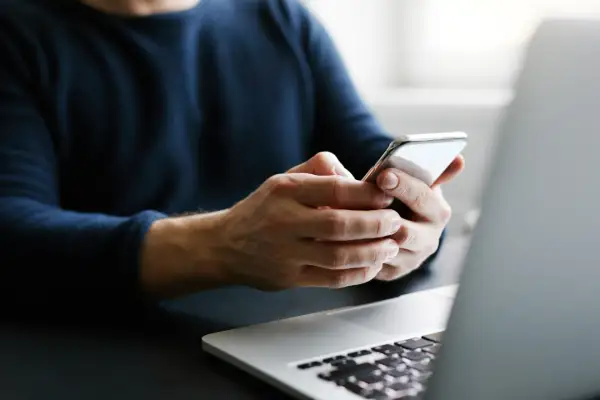Mężczyzna trzyma telefon, szukając informacji, jak przenieść kontakty na kartę SIM. Obok leży laptop.