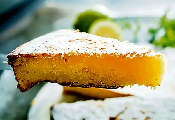 Una fetta di torta caprese al limone, spolverata di zucchero a velo, con fette di limone sullo sfondo.