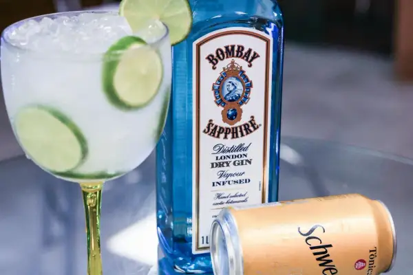 Orzeźwiający drink z tonikiem i limonką, obok butelka ginu Bombay Sapphire i puszka Schweppes. Idealny na letnie wieczory.