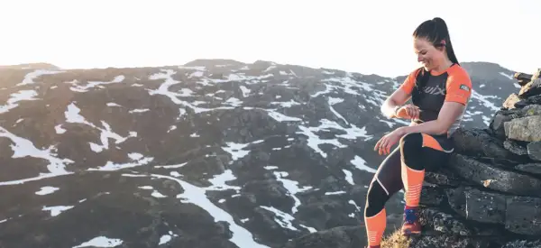 Une femme en tenue de sport s'assoit sur des rochers, regardant sa montre, prête à trouver un trail dans un paysage montagneux enneigé.