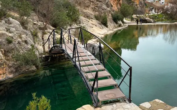 Puente metálico sobre aguas cristalinas, ideal para bolbaite fotos. Rocas y vegetación rodean el estanque.