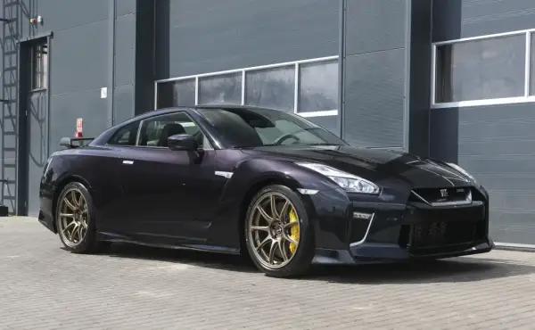 Ciemny Nissan R35 z brązowymi felgami i żółtymi zaciskami hamulcowymi stoi przed szarym budynkiem.