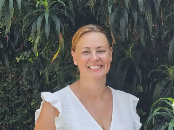 Vrolijke vrouw met blonde haren, lachend in een witte jurk, tegen een achtergrond van weelderig groen. Arjan van Dam zou trots zijn op deze foto.