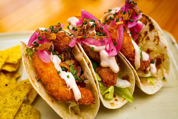 Tres deliciosos tacos de pescado crujientes, cubiertos con cebolla morada encurtida, salsa cremosa y cilantro fresco.