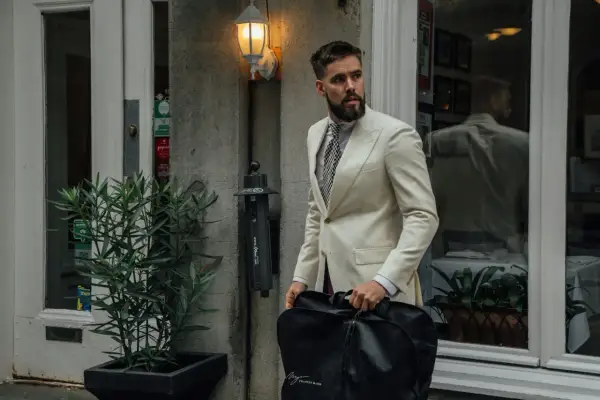 Mann im cremefarbenen Anzug mit gemustertem Hemd und Krawatte, trägt eine schwarze Kleidersacktasche. Ein lässiges Outfit für unterwegs.