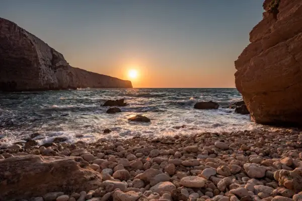 Malta zachód słońca nad kamienistą plażą. Słońce odbija się w falach, a klify tworzą malowniczą scenerię.