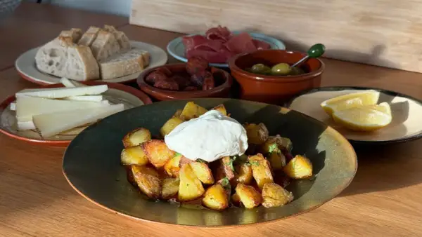 En tallrik med krispiga patatas bravas toppad med en klick krämig sås, omgiven av bröd, ost, chorizo, oliver och citronklyftor.