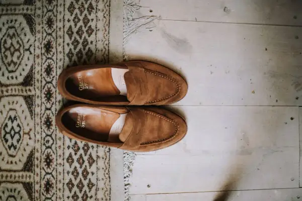 Une paire de mocassins en daim marron, posée sur un tapis à motifs et un parquet. Idéal pour raviver la couleur du daim naturellement.