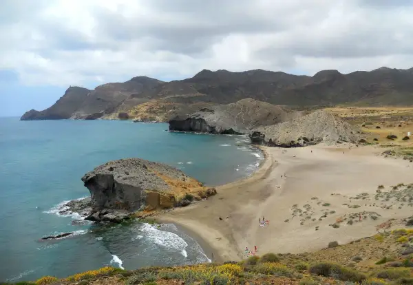 Paisaje de las mejores calas de Cabo de Gata, con formaciones rocosas, arena dorada y mar turquesa bajo un cielo nublado.