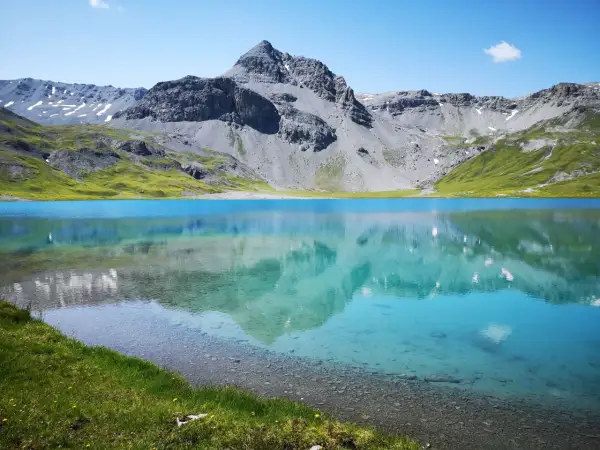 Ein klarer, blauer Bergsee, der **lai da rims**, spiegelt die majestätischen, grauen Berge und grünen Hänge wider.