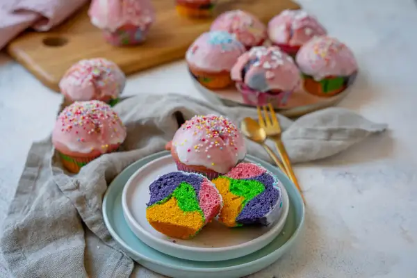 Bunte regenbogen muffins mit rosa Zuckerguss und Streuseln, ein Stück auf einem Teller zeigt das farbenfrohe Innere.