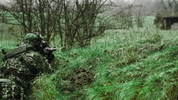 Jugador de airsoft que dispara desde la hierba alta, camuflado y con equipo táctico.
