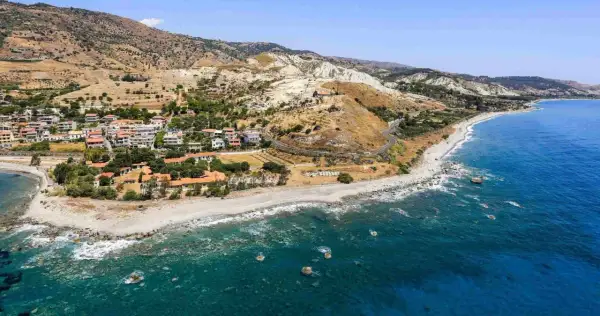 Paesaggio mozzafiato sulla **Calabria Costa Ionica**: case arroccate su colline aride, una spiaggia di ciottoli e il mare blu intenso.