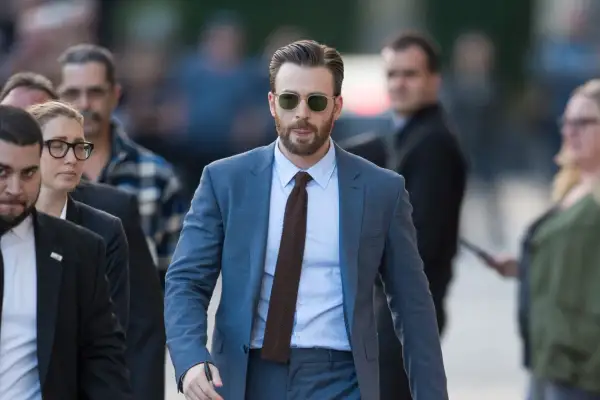 Mann im blauen Anzug, hellblauem Hemd und braunen Schuhen geht mit Sonnenbrille und Bart.