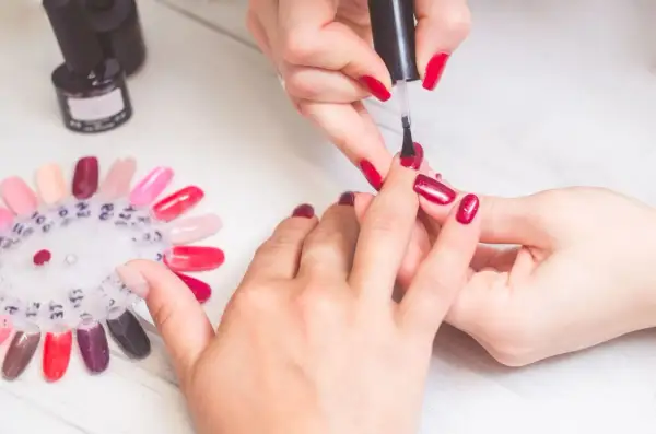 Krok po kroku: jak zrobić manicure. Dłonie w trakcie malowania paznokci na czerwono, obok paleta kolorów lakierów.