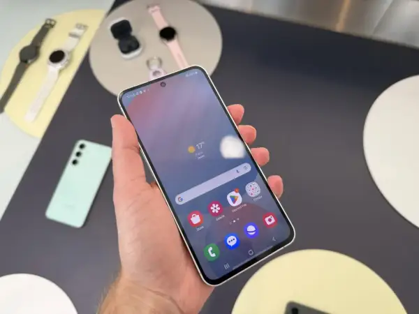 Ręka trzyma smartfon Samsung, pokazując ekran z pogodą i ikonami aplikacji. Obok leżą smartwatch i słuchawki. Czas przed ekranem Samsunga to chwila relaksu.