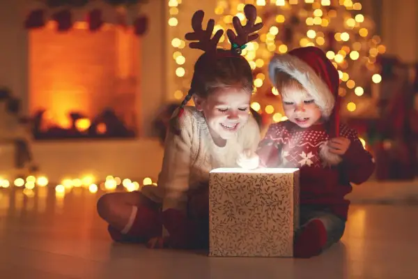 Zwei Kinder mit Rentiergeweih und Weihnachtsmütze öffnen ein leuchtendes Geschenk. Ein magischer Moment im Kindergarten an Weihnachten.