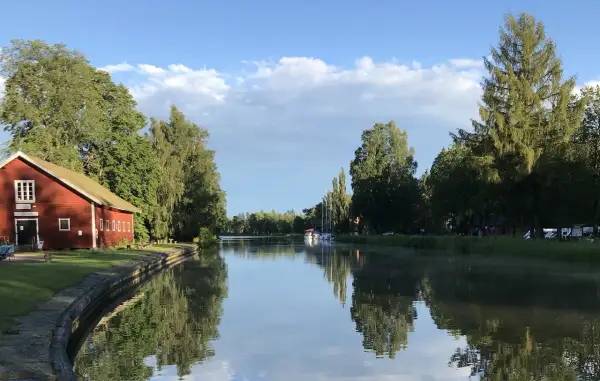 En röd byggnad vid en kanal, troligen Hajstorps slusskafé, med båtar och träd längs vattnet under en blå himmel.