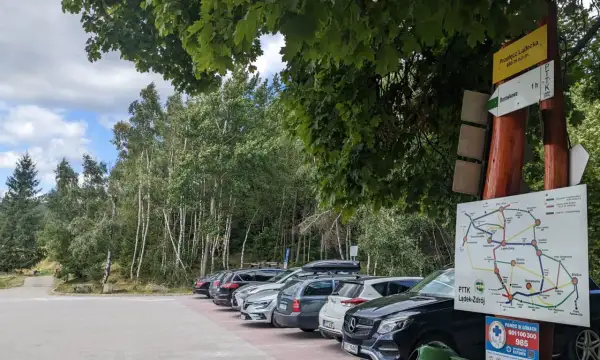 Samochody zaparkowane przy szlaku turystycznym na przełęczy kłodzkiej. W tle las i tablice informacyjne PTTK.