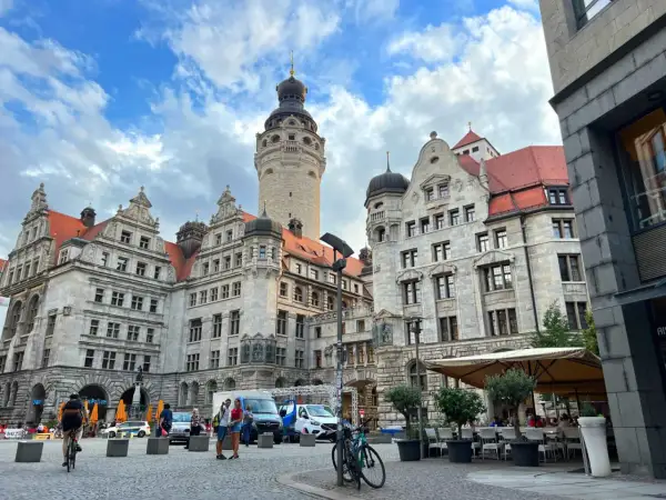 Lipsk trasa: imponujące budynki z wieżą, plac z ludźmi, rowerzystką i kawiarnią pod parasolami.