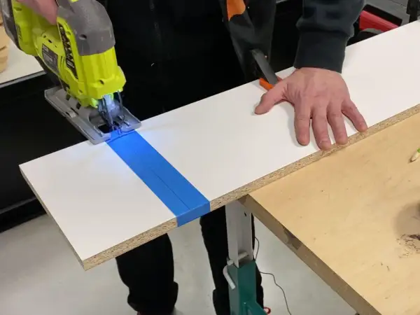 Une scie sauteuse coupe une planche de stratifié, guidée par du ruban bleu. La main d'un bricoleur stabilise la pièce.