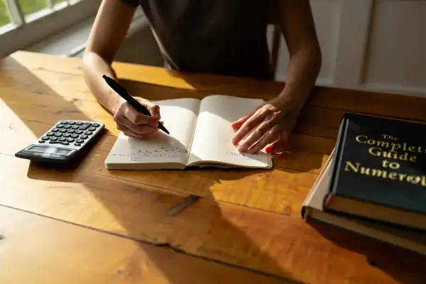 Une personne écrit dans un carnet, une calculatrice et un livre de numérologie à proximité. Cherche-t-elle à trouver une date de naissance gratuite ?