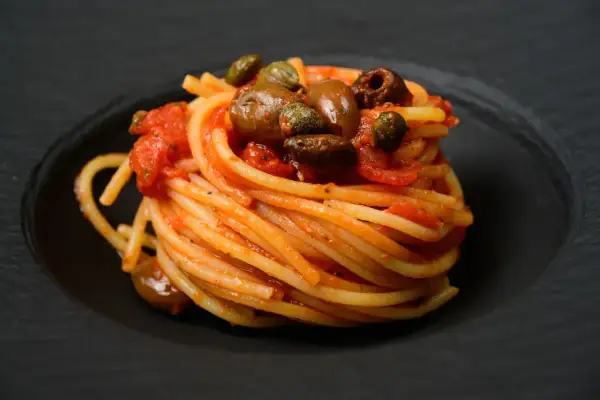 Ein kunstvoll aufgetürmter Teller Spaghetti Puttanesca, garniert mit Oliven und Kapern, auf schwarzem Teller.