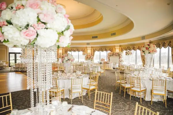 Elegancka sala weselna z okrągłymi stołami, złotymi krzesłami i bogatą dekoracją kwiatową. Stół państwa młodych ozdobiony jest wysokim bukietem róż i białych kwiatów.