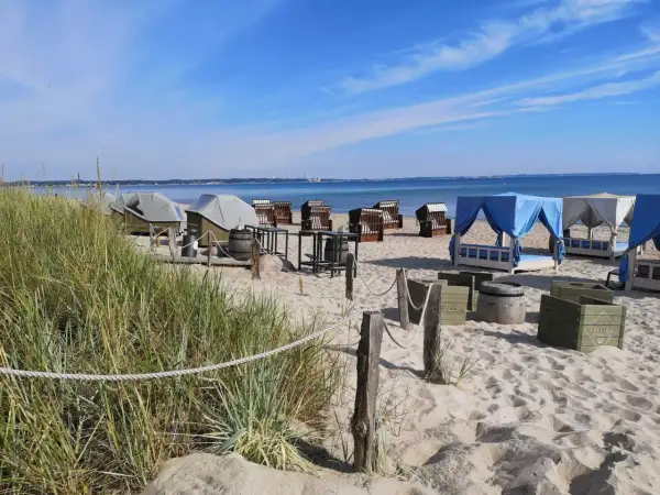 Scharbeutz Strand: Strandkörbe, Liegen unter blauen und weißen Zelten, Tische und Fässer am Sandstrand.