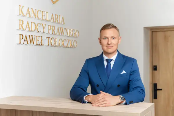 Mężczyzna w niebieskim garniturze za ladą, za nim napis "KANCELaria Radcy Prawnego Paweł Tołoczko".