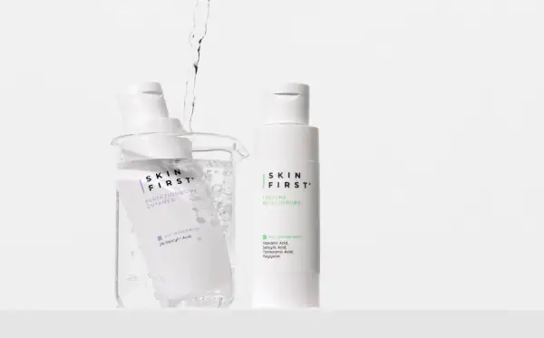 Due flaconi di tonico viso Skin First, uno immerso in un becher con acqua e bollicine, l'altro in piedi.