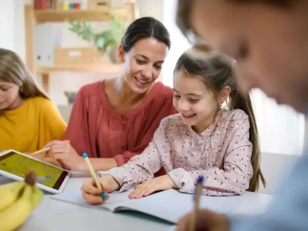 Uśmiechnięta mama pomaga dzieciom w nauce. Jedna córka pisze, druga korzysta z tabletu. To przykład wspólnego uczenia się, gdzie powtarzanie słów, jak w epifora, pomaga zapamiętać.