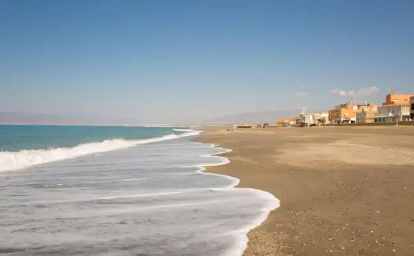 Olas suaves acarician la arena dorada de la playa de Cabo de Gata. Al fondo, un pueblo costero se alza bajo un cielo azul.