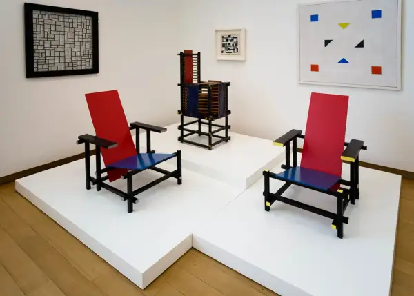 Twee iconische stoelen in rood, blauw en zwart, kenmerkend voor bauhaus kunst, staan naast een abstract schilderij en een sculptuur.