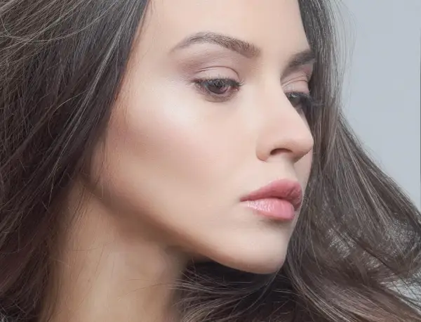 Delikatna twarz kobiety z subtelnym błyskiem na skórze. Makijaż jest tak naturalny, że wygląda jak make up no make up.