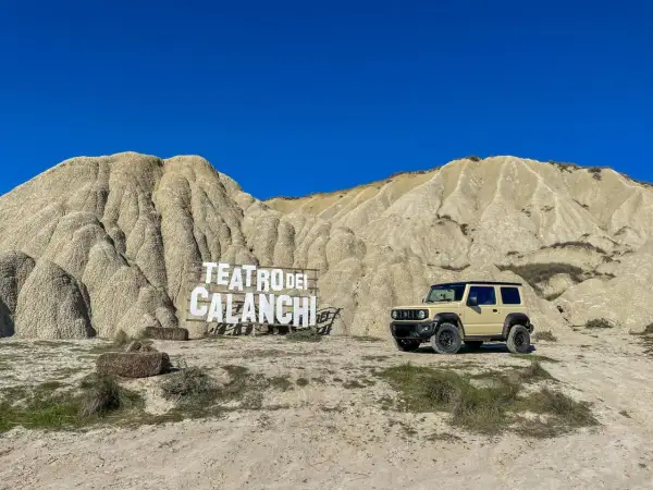 Un fuoristrada beige parcheggiato davanti al **teatro dei calanchi**, un paesaggio lunare con montagne erose e un cartello in legno.