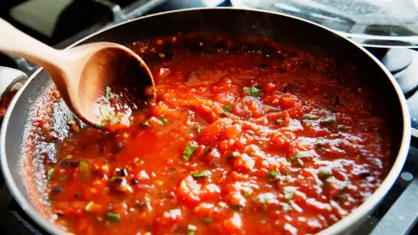 Gęsty, aromatyczny sos do spaghetti gotuje się na patelni. Drewniana łyżka miesza pomidorową masę z ziołami.