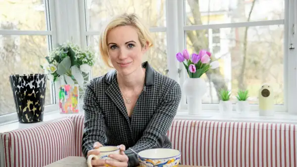 Jina Zachrisson sitter vid ett fönster med en mugg i handen, omgiven av blommor och konst.
