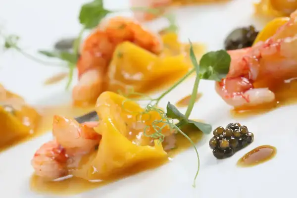 Gamberi succulenti e caviale nero su un letto di pasta con pesce, guarnito con germogli verdi e una salsa dorata.