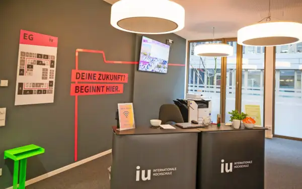 Hier ist die Rezeption der IU Internationale Hochschule. Ein Bildschirm zeigt Bilder, und ein Plan weist den Weg. Was ist die IU? Eine Hochschule, die deine Zukunft beginnt.