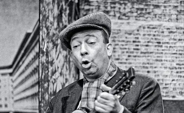 Stanisław Bard Starej Warszawy, w czapce i z gitarą, śpiewa na tle ceglanej ściany.