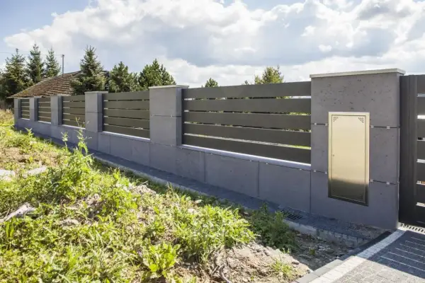 Nowoczesne ogrodzenie z betonowych słupków i ciemnych paneli, wyznaczające nieprzekraczalną linię zabudowy. Złota skrzynka na listy wmurowana w słupek.