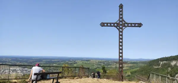 La croix du Dan domine un paysage verdoyant. Deux personnes profitent de la vue depuis un banc.