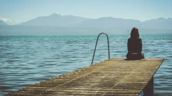 Mujer sentada en un muelle, contemplando el lago y las montañas. Pensamientos de frases de preocuparse por alguien flotan en el aire.