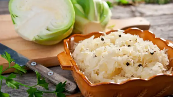 Schüssel mit Sauerkraut, Gewürzen und frischem Weißkohl. Ein Messer und Petersilie deuten auf ein traditionelles Sauerkraut Rezept hin.