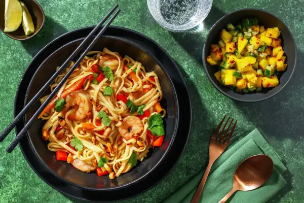 En skål med pad thai med räkor, nudlar, paprika och jordnötter. En liten skål med mangosallad står bredvid.