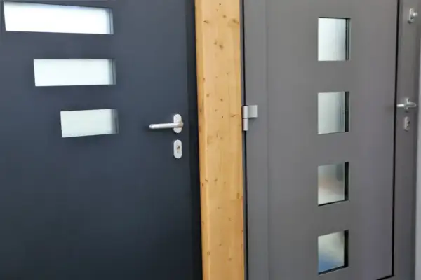Dwie nowoczesne drzwi wejściowe, jedna ciemnoszara z prostokątnymi przeszkleniami, druga szara z kwadratowymi. Dowiedz się, jak uszczelnić drzwi wejściowe.