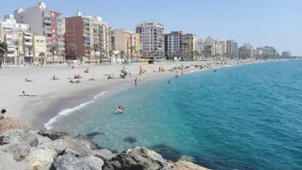 Gente disfrutando del sol y el mar en la playa del Zapillo, con edificios residenciales al fondo.