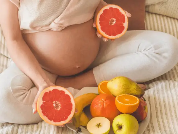 Ciężarna kobieta trzyma połówki grejpfruta, obok talerz z owocami. Czy w ciąży można jeść pomarańcze? Tak, to zdrowe!